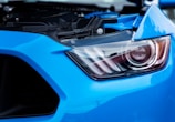 A close-up view of a blue car's headlight, showcasing its sleek design and intricate details. The headlight is modern and features a combination of clear and dark elements under a transparent casing.