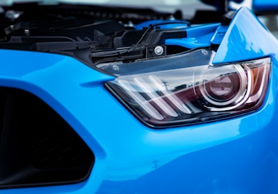 A close-up view of a blue car's headlight, showcasing its sleek design and intricate details. The headlight is modern and features a combination of clear and dark elements under a transparent casing.