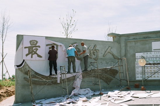 Several people are working on a mural or sign on a concrete wall. They are using scaffolding to reach the upper areas and are applying large pieces of material with Chinese characters on them. The area appears to be outdoors, with a clear sky and some bare trees visible in the background. There are papers and tools scattered on the ground around the workers.