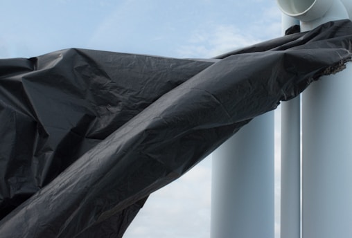 A large black tarp or plastic sheet is wrapped around a structure composed of two white cylindrical pipes. The sky is visible in the background, with light cloud coverage.