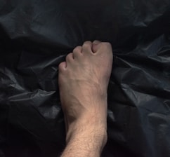 A human foot with visible veins and slight hair growth is placed on a dark, crumpled surface, possibly plastic. The foot is centred with the toes slightly curled.