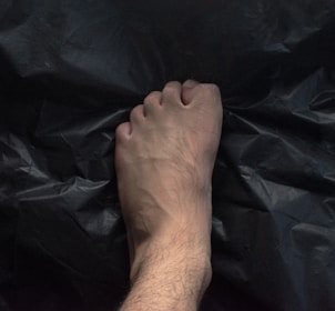 A human foot with visible veins and slight hair growth is placed on a dark, crumpled surface, possibly plastic. The foot is centred with the toes slightly curled.