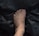 A human foot with visible veins and slight hair growth is placed on a dark, crumpled surface, possibly plastic. The foot is centred with the toes slightly curled.