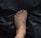 A human foot with visible veins and slight hair growth is placed on a dark, crumpled surface, possibly plastic. The foot is centred with the toes slightly curled.
