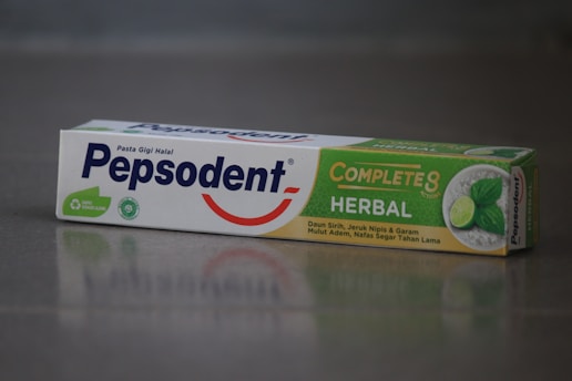 A box of Pepsodent toothpaste with the label 'Complete 8 Herbal' featuring a green and white design. The package includes images of a lime and mint leaves, and contains descriptions in Indonesian about its herbal ingredients. The Pepsodent logo is prominently displayed in blue with a red smile underneath.