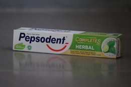 A box of Pepsodent toothpaste with the label 'Complete 8 Herbal' featuring a green and white design. The package includes images of a lime and mint leaves, and contains descriptions in Indonesian about its herbal ingredients. The Pepsodent logo is prominently displayed in blue with a red smile underneath.