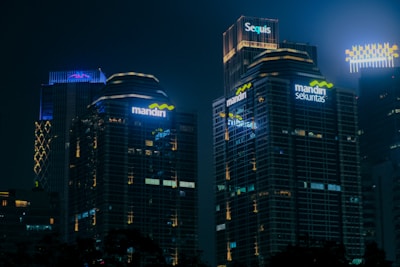 Tall office buildings with illuminated signage stand against a dark night sky. Several of the buildings have glass facades reflecting the lights, and the names 'mandiri', 'mandiri sekuritas', and 'Sequis' are prominently displayed in bright, colored lights. The overall scene depicts a cityscape at night.
