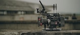 A professional video camera with various attachments, including antennas and a stabilizing rig, is placed on a concrete surface. The background is blurred, suggesting an outdoor or industrial setting.
