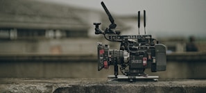 A professional video camera with various attachments, including antennas and a stabilizing rig, is placed on a concrete surface. The background is blurred, suggesting an outdoor or industrial setting.