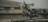 A professional video camera with various attachments, including antennas and a stabilizing rig, is placed on a concrete surface. The background is blurred, suggesting an outdoor or industrial setting.