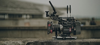 A professional video camera with various attachments, including antennas and a stabilizing rig, is placed on a concrete surface. The background is blurred, suggesting an outdoor or industrial setting.