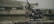 A professional video camera with various attachments, including antennas and a stabilizing rig, is placed on a concrete surface. The background is blurred, suggesting an outdoor or industrial setting.