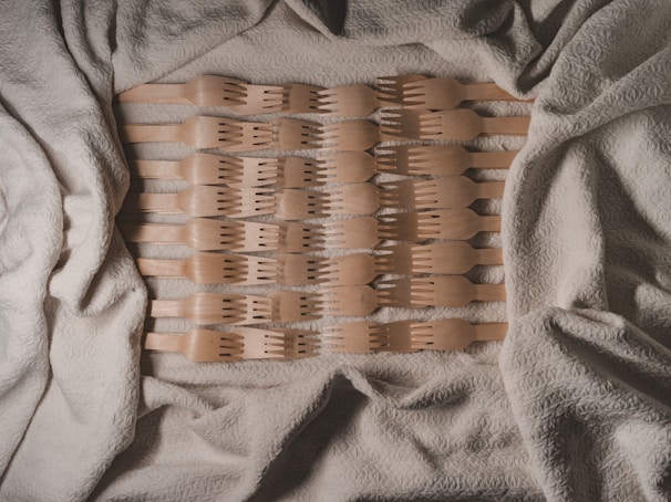 A collection of wooden utensils arranged in a symmetrical pattern on a textured beige fabric background. The utensils are evenly spaced and aligned horizontally, creating a visually pleasing geometric layout.