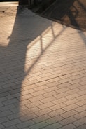 A paved walkway with rectangular tiles arranged in a diagonal pattern. Shadows of a railing and part of a building fall across the surface, creating a blend of light and shadow.