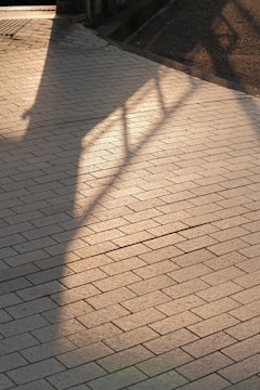 A paved walkway with rectangular tiles arranged in a diagonal pattern. Shadows of a railing and part of a building fall across the surface, creating a blend of light and shadow.