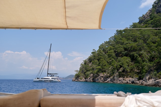 A serene ocean scene with a modern catamaran sailboat anchored near a rocky, tree-covered coastline. The foreground includes a shaded area and comfortable seating with a cloth canopy overhead, suggesting a viewpoint from another boat. The water is calm and clear, reflecting the blue sky with a few clouds.
