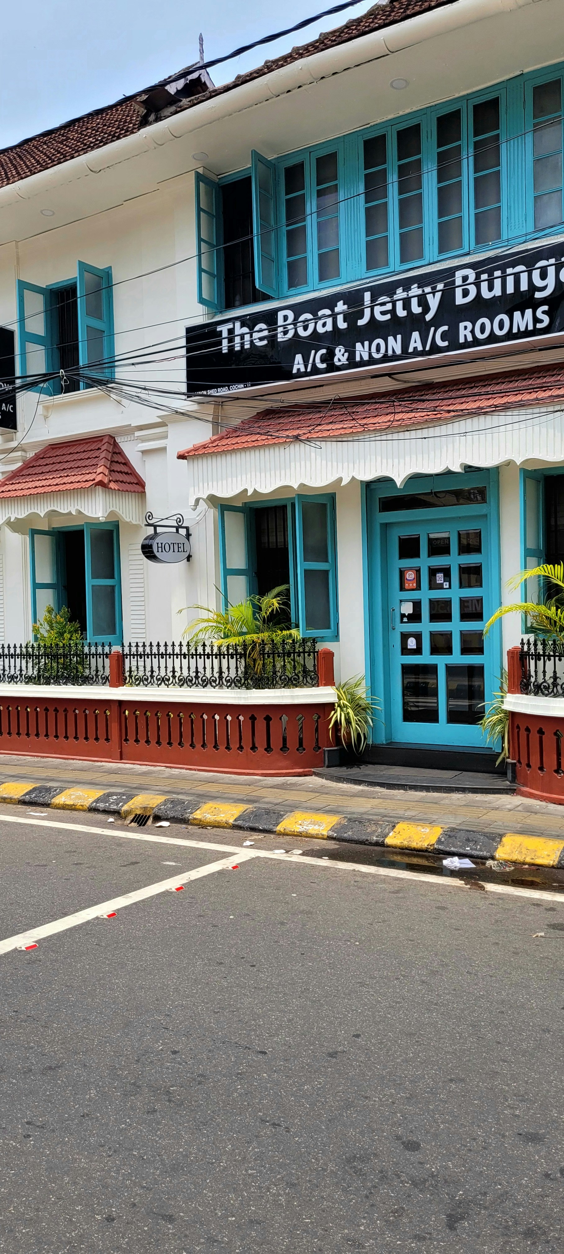 A charming two-story building with teal window frames and doors set against a white facade. The brown roof complements the neatly trimmed plants that line the patio. A street runs in front with a zebra crossing and yellow-black striped curbs. A signboard advertises the availability of both air-conditioned and non-air-conditioned rooms.