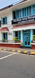 A charming two-story building with teal window frames and doors set against a white facade. The brown roof complements the neatly trimmed plants that line the patio. A street runs in front with a zebra crossing and yellow-black striped curbs. A signboard advertises the availability of both air-conditioned and non-air-conditioned rooms.