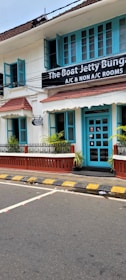 A charming two-story building with teal window frames and doors set against a white facade. The brown roof complements the neatly trimmed plants that line the patio. A street runs in front with a zebra crossing and yellow-black striped curbs. A signboard advertises the availability of both air-conditioned and non-air-conditioned rooms.