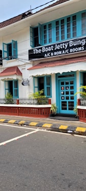 A charming two-story building with teal window frames and doors set against a white facade. The brown roof complements the neatly trimmed plants that line the patio. A street runs in front with a zebra crossing and yellow-black striped curbs. A signboard advertises the availability of both air-conditioned and non-air-conditioned rooms.
