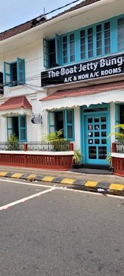A charming two-story building with teal window frames and doors set against a white facade. The brown roof complements the neatly trimmed plants that line the patio. A street runs in front with a zebra crossing and yellow-black striped curbs. A signboard advertises the availability of both air-conditioned and non-air-conditioned rooms.