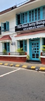 A charming two-story building with teal window frames and doors set against a white facade. The brown roof complements the neatly trimmed plants that line the patio. A street runs in front with a zebra crossing and yellow-black striped curbs. A signboard advertises the availability of both air-conditioned and non-air-conditioned rooms.