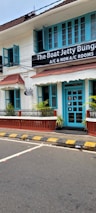 A charming two-story building with teal window frames and doors set against a white facade. The brown roof complements the neatly trimmed plants that line the patio. A street runs in front with a zebra crossing and yellow-black striped curbs. A signboard advertises the availability of both air-conditioned and non-air-conditioned rooms.