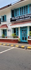 A charming two-story building with teal window frames and doors set against a white facade. The brown roof complements the neatly trimmed plants that line the patio. A street runs in front with a zebra crossing and yellow-black striped curbs. A signboard advertises the availability of both air-conditioned and non-air-conditioned rooms.