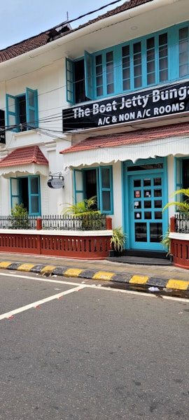 A charming two-story building with teal window frames and doors set against a white facade. The brown roof complements the neatly trimmed plants that line the patio. A street runs in front with a zebra crossing and yellow-black striped curbs. A signboard advertises the availability of both air-conditioned and non-air-conditioned rooms.