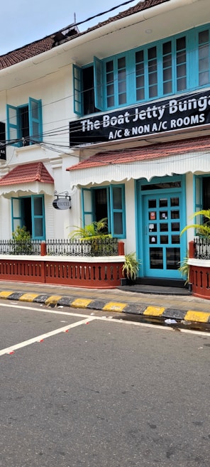 A charming two-story building with teal window frames and doors set against a white facade. The brown roof complements the neatly trimmed plants that line the patio. A street runs in front with a zebra crossing and yellow-black striped curbs. A signboard advertises the availability of both air-conditioned and non-air-conditioned rooms.