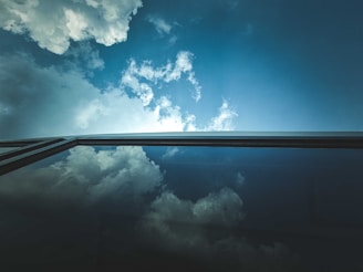 A clear blue sky with white clouds is reflected on the surface of a large glass window, creating a seamless continuation between the sky and its reflection.
