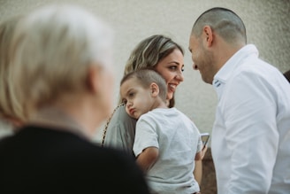 A group of people gathered together, with a focus on a woman smiling while holding a young child who looks tired. Another adult stands close by, facing them with a cheerful expression. The background is blurred, highlighting the interactions among the individuals.