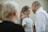 A group of people gathered together, with a focus on a woman smiling while holding a young child who looks tired. Another adult stands close by, facing them with a cheerful expression. The background is blurred, highlighting the interactions among the individuals.