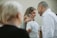 A group of people gathered together, with a focus on a woman smiling while holding a young child who looks tired. Another adult stands close by, facing them with a cheerful expression. The background is blurred, highlighting the interactions among the individuals.