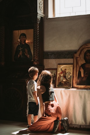 A woman and a child are kneeling and gazing at religious icons in a dimly lit room. Sunlight streams in through a window, casting soft light on the icons displayed on a table. The room has ornate wooden furniture and decorative elements.