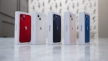 Five iPhone boxes are lined up on a marble surface. Each box showcases a different color of the iPhone, including red, white, black, pink, and blue. The background features a softly blurred light pattern.