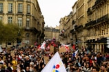 A large crowd of people participates in a protest or demonstration on a city street lined with historical buildings. Many individuals are holding signs and banners, some with red flags, while others carry placards with text. The scene exudes a sense of communal action amidst an urban backdrop with trees and old architecture.