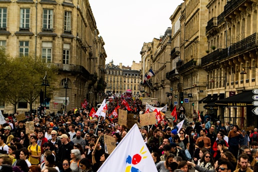 A large crowd of people participates in a protest or demonstration on a city street lined with historical buildings. Many individuals are holding signs and banners, some with red flags, while others carry placards with text. The scene exudes a sense of communal action amidst an urban backdrop with trees and old architecture.