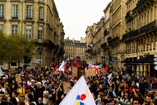 A large crowd of people participates in a protest or demonstration on a city street lined with historical buildings. Many individuals are holding signs and banners, some with red flags, while others carry placards with text. The scene exudes a sense of communal action amidst an urban backdrop with trees and old architecture.