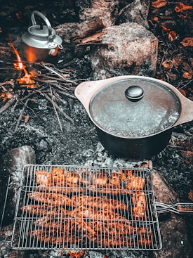 A campfire scene featuring a grill rack filled with cooked sausages above glowing embers. Nearby, a metal pot with a lid is placed on the ashes, and a kettle sits to the left. Surrounding the fire are rocks and some scattered branches.