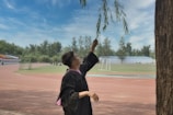 A person in a graduation gown reaches up to touch the branches of a tree, standing on a sports track surrounded by greenery. In the background, there is a field with a goalpost and several other individuals gathered in the distance. The sky is partly cloudy, creating a serene atmosphere.
