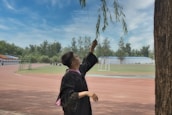 A person in a graduation gown reaches up to touch the branches of a tree, standing on a sports track surrounded by greenery. In the background, there is a field with a goalpost and several other individuals gathered in the distance. The sky is partly cloudy, creating a serene atmosphere.