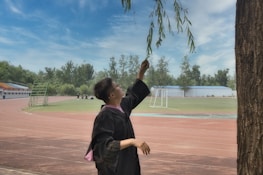 A person in a graduation gown reaches up to touch the branches of a tree, standing on a sports track surrounded by greenery. In the background, there is a field with a goalpost and several other individuals gathered in the distance. The sky is partly cloudy, creating a serene atmosphere.