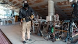 A person wearing protective clothing and a welding mask is working in a workshop or studio. There are various tools and pieces of equipment scattered around, including a welding machine and some metal structures. The space appears cluttered and is somewhat industrial in appearance, with an unfinished ceiling and assorted materials lying around.