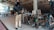 A person wearing protective clothing and a welding mask is working in a workshop or studio. There are various tools and pieces of equipment scattered around, including a welding machine and some metal structures. The space appears cluttered and is somewhat industrial in appearance, with an unfinished ceiling and assorted materials lying around.
