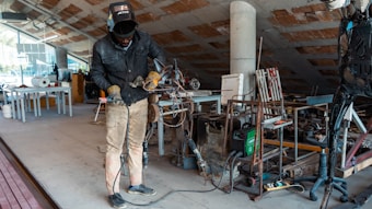 A person wearing protective clothing and a welding mask is working in a workshop or studio. There are various tools and pieces of equipment scattered around, including a welding machine and some metal structures. The space appears cluttered and is somewhat industrial in appearance, with an unfinished ceiling and assorted materials lying around.