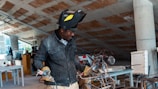 A person wearing a welding helmet and gloves is working on a metal object in an industrial or workshop setting. There are various tools and pieces of equipment scattered around the room, including tables, wires, and machinery. The environment appears to be cluttered with construction or fabrication materials.
