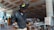 A person wearing a welding helmet and gloves is working on a metal object in an industrial or workshop setting. There are various tools and pieces of equipment scattered around the room, including tables, wires, and machinery. The environment appears to be cluttered with construction or fabrication materials.