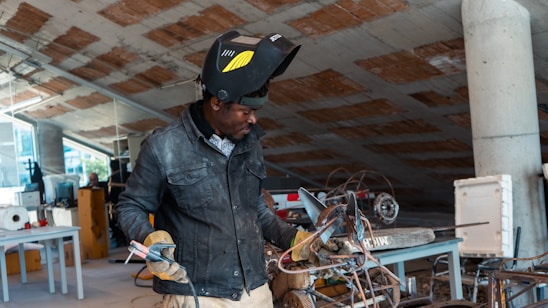 A person wearing a welding helmet and gloves is working on a metal object in an industrial or workshop setting. There are various tools and pieces of equipment scattered around the room, including tables, wires, and machinery. The environment appears to be cluttered with construction or fabrication materials.