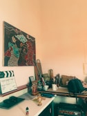 A cluttered workspace features a wall adorned with a colorful painting depicting fantastical figures and clouds. The desk below holds various items including a clapperboard with scene details, a decorative bottle, a small toy figure, a teacup, and an assorted collection of pens and tape. In the background, a couple of badminton rackets are propped up, alongside a backpack and some paper bags. An amplifier sits under the desk.